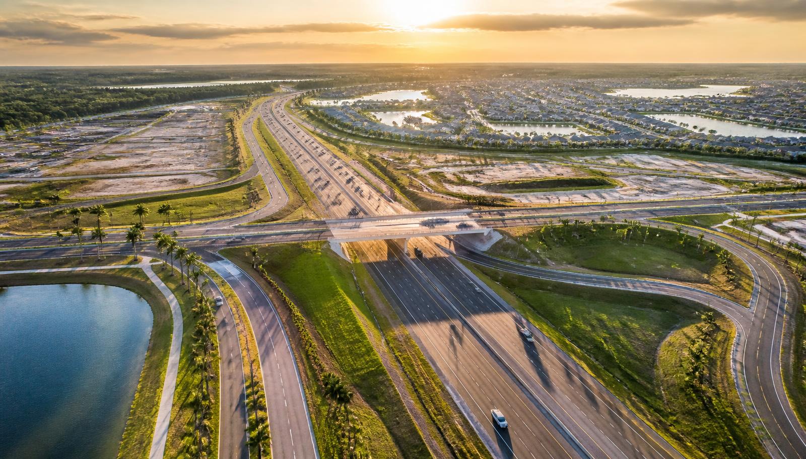 Aerial golden-hour view of an interstate interchange in northern Wesley Chapel, Florida, with palm-lined frontage roads, cleared development parcels and master-planned neighborhoods with lakes in the distance